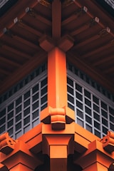 The image shows the detailed architectural design of a traditional Japanese-style structure, featuring vibrant orange wooden beams and intricate fittings. The design emphasizes symmetry and craftsmanship, with a grid-like window section beneath the roof.