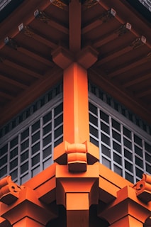 The image shows the detailed architectural design of a traditional Japanese-style structure, featuring vibrant orange wooden beams and intricate fittings. The design emphasizes symmetry and craftsmanship, with a grid-like window section beneath the roof.