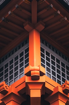 The image shows the detailed architectural design of a traditional Japanese-style structure, featuring vibrant orange wooden beams and intricate fittings. The design emphasizes symmetry and craftsmanship, with a grid-like window section beneath the roof.