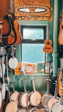 A room filled with musical instruments, predominantly banjos and guitars, arranged on a floor and hung on walls. There is a window with light streaming in, providing a softly lit atmosphere. Various posters and memorabilia add a vintage charm to the setting.