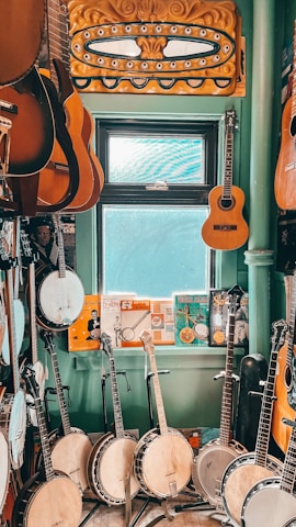 A room filled with musical instruments, predominantly banjos and guitars, arranged on a floor and hung on walls. There is a window with light streaming in, providing a softly lit atmosphere. Various posters and memorabilia add a vintage charm to the setting.