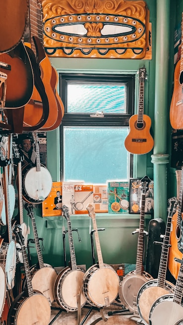 A room filled with musical instruments, predominantly banjos and guitars, arranged on a floor and hung on walls. There is a window with light streaming in, providing a softly lit atmosphere. Various posters and memorabilia add a vintage charm to the setting.