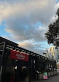 An outdoor scene featuring a building labeled 'The General Assembly' with a prominent red sign. The structure is surrounded by potted plants, and tall modern skyscrapers are visible in the background. The sky is partly cloudy with patches of blue.