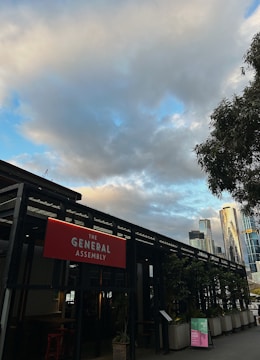 An outdoor scene featuring a building labeled 'The General Assembly' with a prominent red sign. The structure is surrounded by potted plants, and tall modern skyscrapers are visible in the background. The sky is partly cloudy with patches of blue.