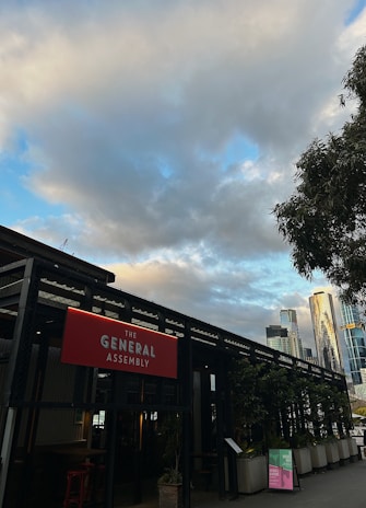 An outdoor scene featuring a building labeled 'The General Assembly' with a prominent red sign. The structure is surrounded by potted plants, and tall modern skyscrapers are visible in the background. The sky is partly cloudy with patches of blue.