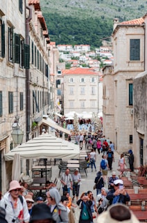 A bustling street scene in a historic town, featuring people walking along a narrow stone-paved path flanked by old stone buildings with green shutters. Numerous tourists are visible, some taking photos and others gathering in small groups. Several large umbrellas provide shade over outdoor seating areas. In the background, a hillside filled with houses can be seen under a cloudy sky.