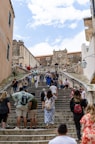 A busy stone staircase with tourists climbing and descending. The architecture is historic, featuring a large stone structure with a clock at the top. Several people are gathered on the steps, some seated and others walking. The scene suggests a vibrant tourist destination.