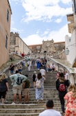 A busy stone staircase with tourists climbing and descending. The architecture is historic, featuring a large stone structure with a clock at the top. Several people are gathered on the steps, some seated and others walking. The scene suggests a vibrant tourist destination.