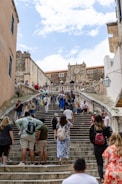 A busy stone staircase with tourists climbing and descending. The architecture is historic, featuring a large stone structure with a clock at the top. Several people are gathered on the steps, some seated and others walking. The scene suggests a vibrant tourist destination.