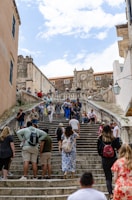 A busy stone staircase with tourists climbing and descending. The architecture is historic, featuring a large stone structure with a clock at the top. Several people are gathered on the steps, some seated and others walking. The scene suggests a vibrant tourist destination.