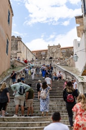 A busy stone staircase with tourists climbing and descending. The architecture is historic, featuring a large stone structure with a clock at the top. Several people are gathered on the steps, some seated and others walking. The scene suggests a vibrant tourist destination.