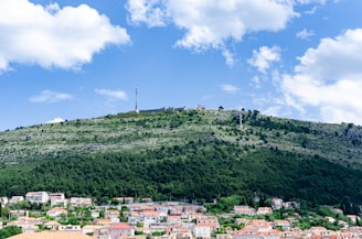 A scenic view of a hillside with lush greenery and a few scattered buildings. A prominent radio tower stands at the top of the hill, with a clear blue sky and fluffy white clouds overhead. Below the hill, a residential area with houses featuring orange roofs lies in the foreground.