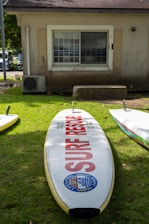 A surf rescue board with a logo and text 'surf rescue' lies on a grassy area in front of a small building with a window and an air conditioning unit. Shadows from nearby trees partially cover the grass.