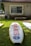 A surf rescue board with a logo and text 'surf rescue' lies on a grassy area in front of a small building with a window and an air conditioning unit. Shadows from nearby trees partially cover the grass.