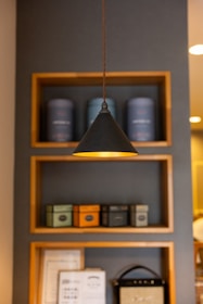 A stylish interior setup featuring a hanging pendant light against a gray wall with wooden shelves. The shelves contain various decorative containers, with some resembling vintage tins, and framed items below them. The focus is on the modern lighting accentuated by a warm glow.