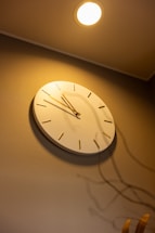 A wall clock with a minimalistic design and white background hangs on a wall. The clock features simple gold hour markers and hands. A bright ceiling light illuminates the scene, casting shadows of decorative objects below the clock.