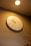 A wall clock with a minimalistic design and white background hangs on a wall. The clock features simple gold hour markers and hands. A bright ceiling light illuminates the scene, casting shadows of decorative objects below the clock.