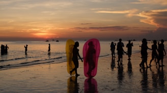 Silhouettes of people walking along a beach during sunset with vibrant colors in the sky. The scene captures families and groups enjoying the water, including two children holding colorful inflatable floaties.