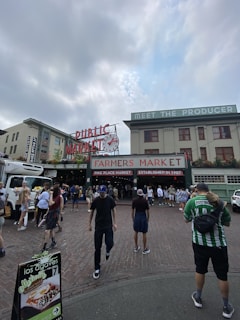 A bustling outdoor market scene with a large sign reading 'Farmers Market' and 'Public Market' located in an urban setting. Several people are walking on the cobblestone road, some carrying bags, and others holding flowers. A produce truck is parked on the side while market stalls are visible in the background.