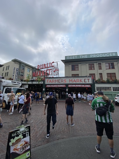 A bustling outdoor market scene with a large sign reading 'Farmers Market' and 'Public Market' located in an urban setting. Several people are walking on the cobblestone road, some carrying bags, and others holding flowers. A produce truck is parked on the side while market stalls are visible in the background.
