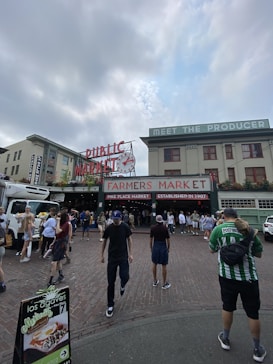 A bustling outdoor market scene with a large sign reading 'Farmers Market' and 'Public Market' located in an urban setting. Several people are walking on the cobblestone road, some carrying bags, and others holding flowers. A produce truck is parked on the side while market stalls are visible in the background.
