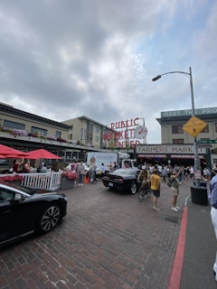 A bustling outdoor market scene featuring a large 'Public Market Center' sign with a clock. People are walking around, some with bikes, near stalls with red umbrellas and flower arrangements. Cars are parked along a cobblestone street, and the sky is partly cloudy.
