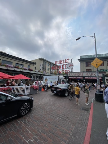 A bustling outdoor market scene featuring a large 'Public Market Center' sign with a clock. People are walking around, some with bikes, near stalls with red umbrellas and flower arrangements. Cars are parked along a cobblestone street, and the sky is partly cloudy.