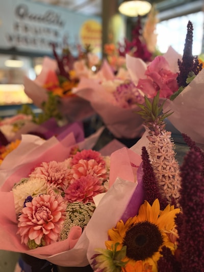 A vibrant display of floral bouquets featuring a variety of brightly colored flowers. The arrangements include sunflowers, dahlias, and delicate lavender spikes, all wrapped in pastel-colored paper. The scene is set in what appears to be a flower shop with a blurred and colorful background.