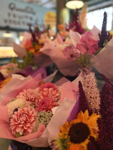 A vibrant display of floral bouquets featuring a variety of brightly colored flowers. The arrangements include sunflowers, dahlias, and delicate lavender spikes, all wrapped in pastel-colored paper. The scene is set in what appears to be a flower shop with a blurred and colorful background.