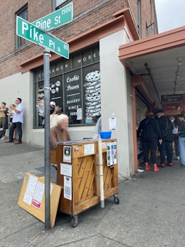A person is seated at a piano on a city street corner near the intersection of Pike Place and Pine Street. The backdrop features a bakery with a large cinnamon roll image in the window. Several pedestrians are visible, some waiting in line and others engaged in conversation, one holding a bouquet of flowers. The overall scene has an urban atmosphere.