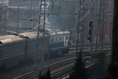 A train is moving along a network of railway tracks surrounded by a complex array of overhead wires and industrial structures. The train consists of several cars, with a smoke-emitting engine at the front, traveling through an urban industrial area. The scene is framed by metal towers and signals, with buildings visible in the background.