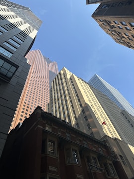 Several tall buildings of varying architectural styles converge upwards towards a clear blue sky. The buildings range in colors from brown-red brick to beige and gray tones, creating a contrast with the bright sky.