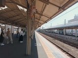A sunlit train station platform with a few people waiting. Some individuals are carrying bags and luggage, suggesting they might be travelers. The platform is covered with a large roof, and the tracks are visible running parallel to the platform.
