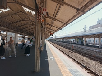 A sunlit train station platform with a few people waiting. Some individuals are carrying bags and luggage, suggesting they might be travelers. The platform is covered with a large roof, and the tracks are visible running parallel to the platform.