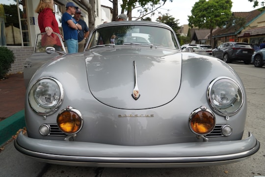 A classic silver Porsche car is parked on a street with a group of people gathering around it. The vintage vehicle features round headlights and an elegant design, showcasing its timeless appeal. In the background, there are trees and buildings lining the street.