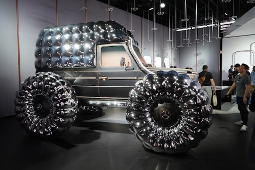 An unusual, futuristic vehicle with a shiny, metallic surface and large, exaggerated tires. The design features a bubble-like texture on both the main body and the wheels. The vehicle is displayed indoors, with people observing it around the showroom.