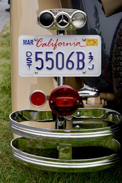 A close-up view of the rear end of a vintage car, featuring a California license plate with the number 5506B. The plate is mounted above a red circular taillight. Shiny chrome details and a distinct car emblem add to the classic car aesthetic. The car is parked on a grassy area.