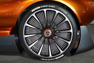 Close-up of a car wheel with a futuristic and intricate design, featuring a large number of metal spokes and a prominent Mercedes-Benz logo in the center. The surrounding car body is a metallic orange color, enhancing the modern aesthetic.