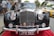 A vintage black automobile with a shiny chrome grille parked on a red carpet. The front view displays multiple decorative badges and a license plate reading 'JYW 36D'. In the background, a crowd of people stand near a yellow sports car.