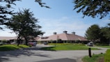 A large building with a sloped, reddish-brown roof is surrounded by well-maintained lawns and tall trees. Several vehicles are parked near the entrance, and a few people can be seen walking nearby. The sky is clear with a few clouds, and the scene is well-lit, suggesting daytime.