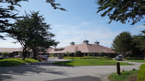 A large building with a sloped, reddish-brown roof is surrounded by well-maintained lawns and tall trees. Several vehicles are parked near the entrance, and a few people can be seen walking nearby. The sky is clear with a few clouds, and the scene is well-lit, suggesting daytime.