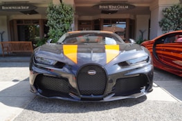 A high-end sports car with a black and orange color scheme is parked in front of a resort shop and a wine cellar. The car has a sleek design with prominent front grilles and headlights. There is another car with a matching orange theme partially visible on the right side. The environment includes some green plants and wooden benches.