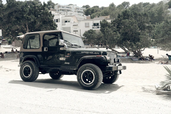 A black Jeep Wrangler is parked on a sandy area near a beach, surrounded by trees and people relaxing under the shade. In the background, white buildings with multiple stories can be seen nestled among the trees.