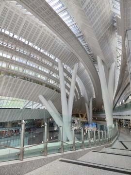 A modern architectural indoor space featuring large, curved, white beams and a high, expansive ceiling with numerous skylights. There is a sleek glass railing along a walkway, and below it, multiple levels can be seen with seating areas and small groups of people.