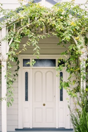 A white front door surrounded by a wooden arch and vine plants, with some greenery and small flowers cascading around the entrance. The façade is made of light-colored wooden siding with panels beside the door that feature textured glass.