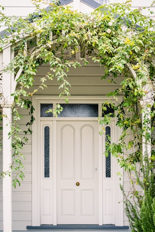 A white front door surrounded by a wooden arch and vine plants, with some greenery and small flowers cascading around the entrance. The fa&ccedil;ade is made of light-colored wooden siding with panels beside the door that feature textured glass.