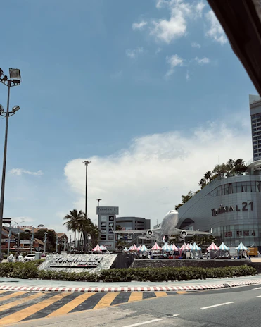 A commercial area featuring a large airplane model displayed prominently in front of a shopping center called Terminal 21. There are several colorful tents set up in front of the complex, alongside palm trees. The sky is clear with a few clouds, and there is a road with directional markings in the foreground.