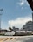 A commercial area featuring a large airplane model displayed prominently in front of a shopping center called Terminal 21. There are several colorful tents set up in front of the complex, alongside palm trees. The sky is clear with a few clouds, and there is a road with directional markings in the foreground.