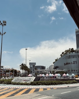 A commercial area featuring a large airplane model displayed prominently in front of a shopping center called Terminal 21. There are several colorful tents set up in front of the complex, alongside palm trees. The sky is clear with a few clouds, and there is a road with directional markings in the foreground.
