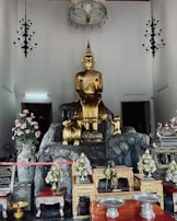 A large golden Buddha statue is the central focus, placed on a rocky pedestal. Surrounding the statue are various items, including ornate vases with pink and white flowers, smaller statues, incense holders, and decorative tables with intricate designs. The room features a high ceiling with hanging chandeliers and lamps, along with framed pictures or artworks on the walls.